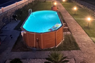 Piscina in acciaio fuori terra ABAK effetto legno Osmose 760x395x132, installata in giardino con suggestiva illuminazione serale. Una soluzione elegante e resistente per chi cerca una piscina da esterno facile da montare, robusta e dal design moderno.